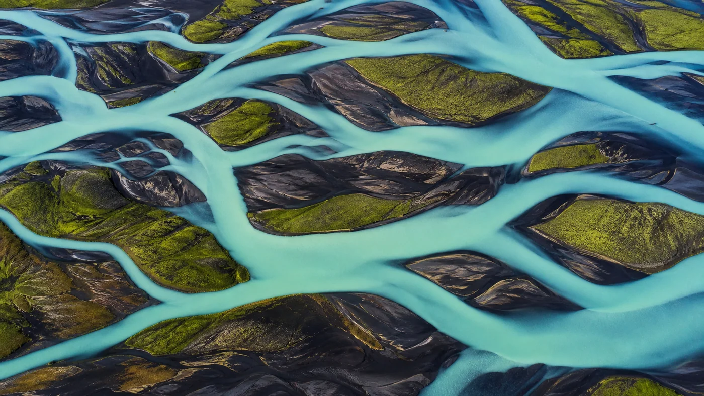 Glacial River Braids — Turquoise channels through black sand and green moss become real aerial abstract art.